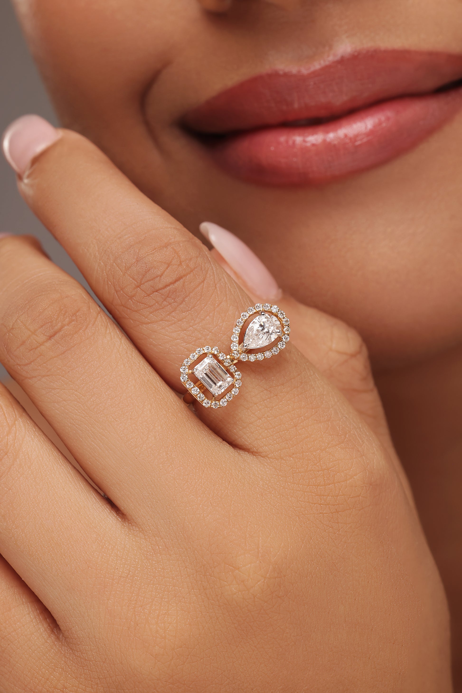 Close-up shot of a hand modeling a toi et moi ring with lab grown diamond in it, featuring a pear-shaped diamond and an emerald-cut diamond, each surrounded by a round halo.