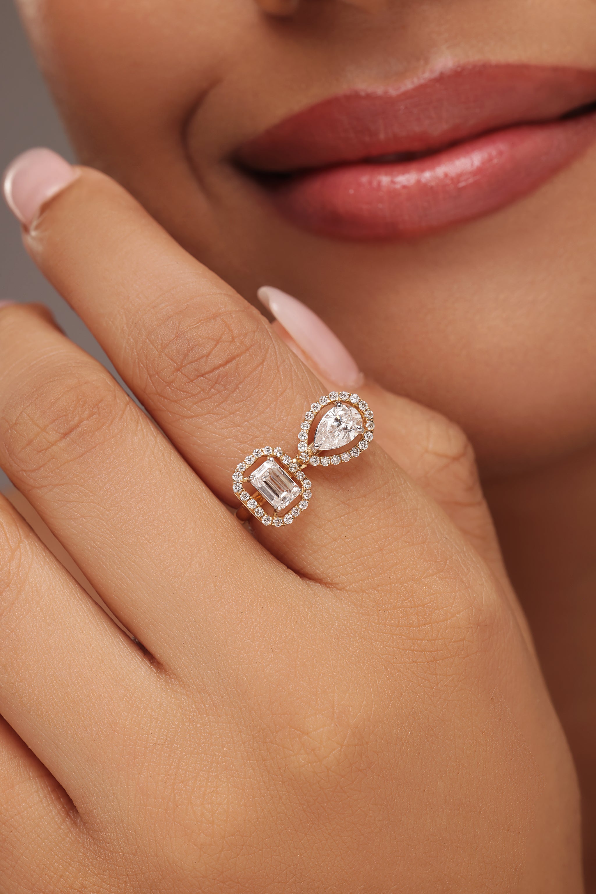 Close-up shot of a hand modeling a toi et moi ring with lab grown diamond in it, featuring a pear-shaped diamond and an emerald-cut diamond, each surrounded by a round halo.