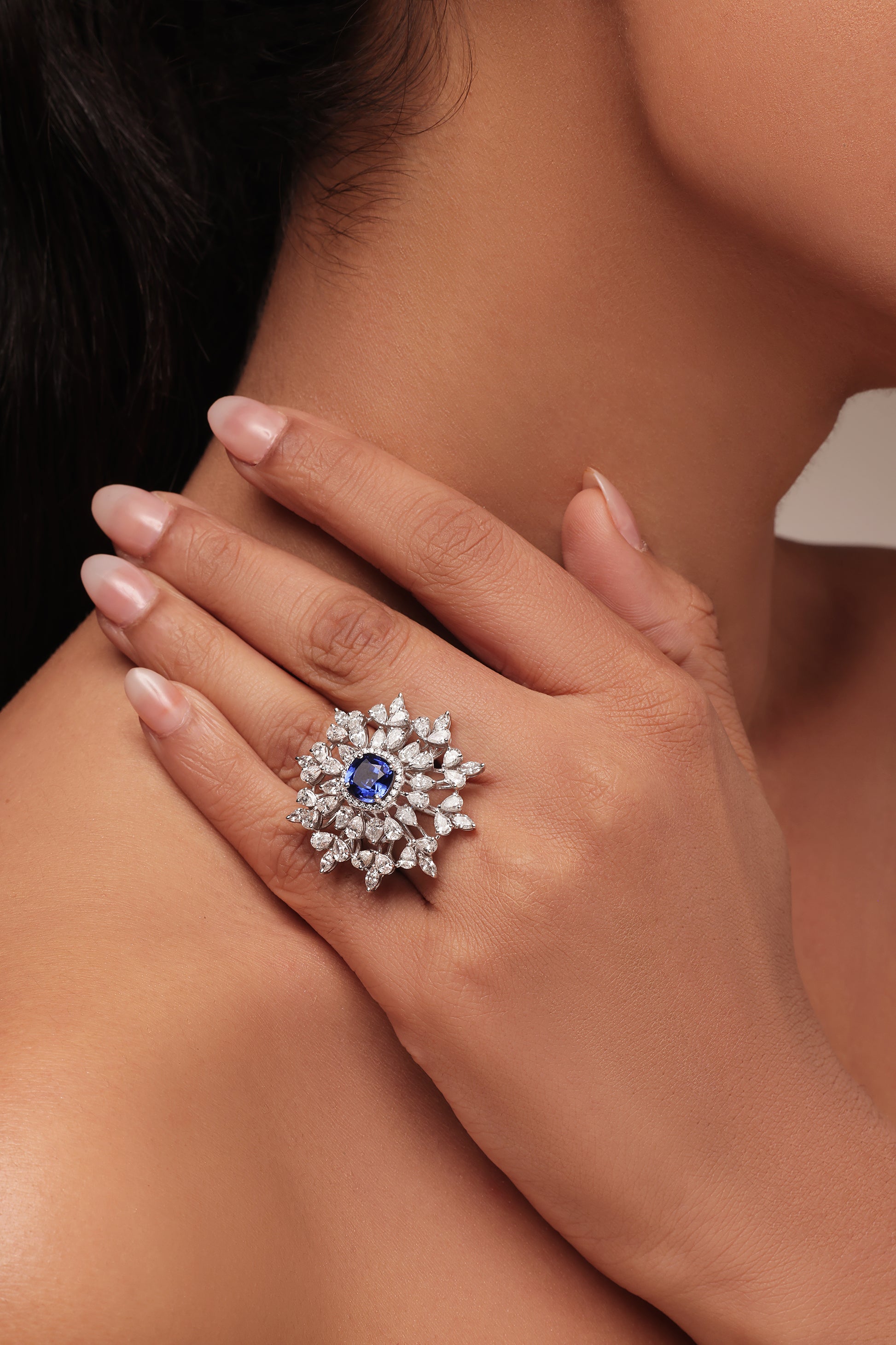 Close-up shot of a hand modeling a large cluster ring with lab grown diamond in it, featuring a central round blue stone surrounded by a dazzling burst of clear marquise and pear diamond accent stones.