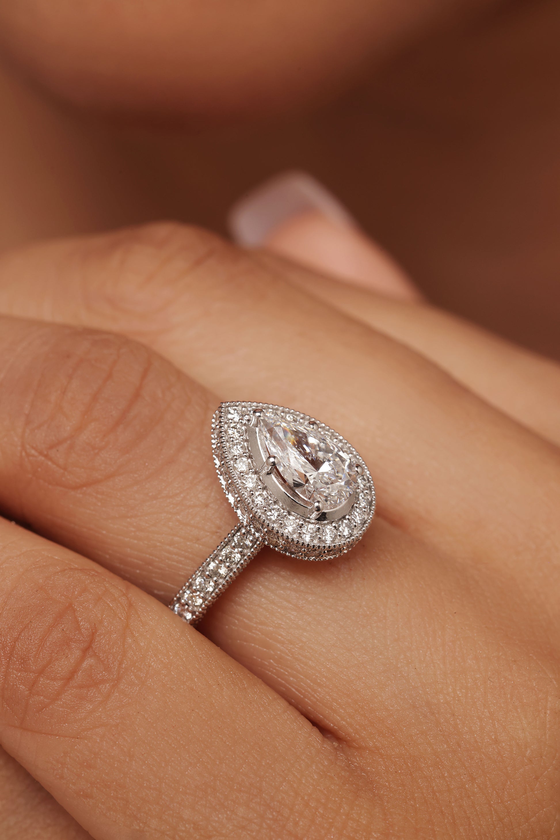 Close-up shot of a hand modeling an engagement-style ring with a pear diamond center stone, surrounded by a halo and set on a round diamond pave band, featuring lab grown diamond in it.