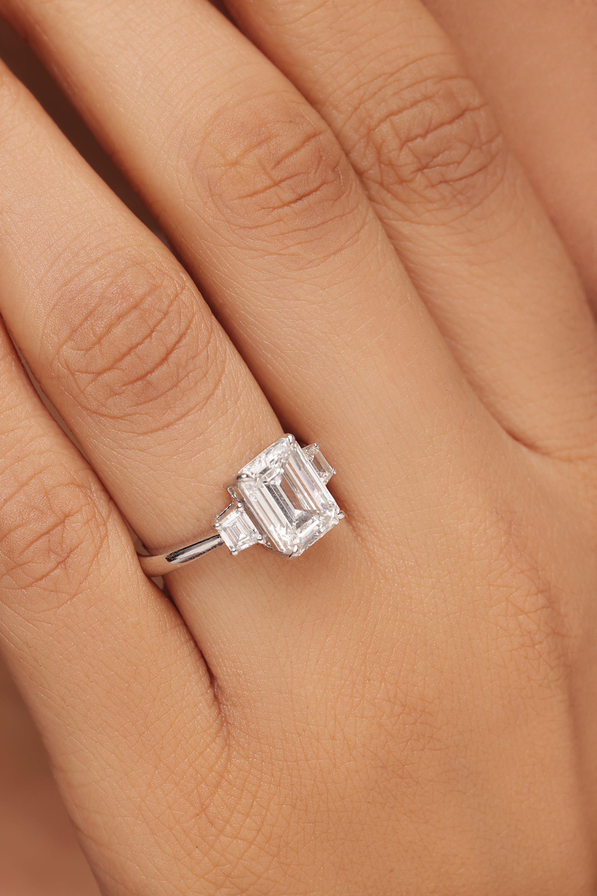 Close-up of a woman's hand showcasing a classic three-stone lab diamond engagement ring set in white gold or platinum. The ring features a rectangular emerald cut center stone flanked by two smaller baguette or step-cut diamonds. Ideal for promoting vintage-style ethical fine jewellery and high-clarity diamond rings.