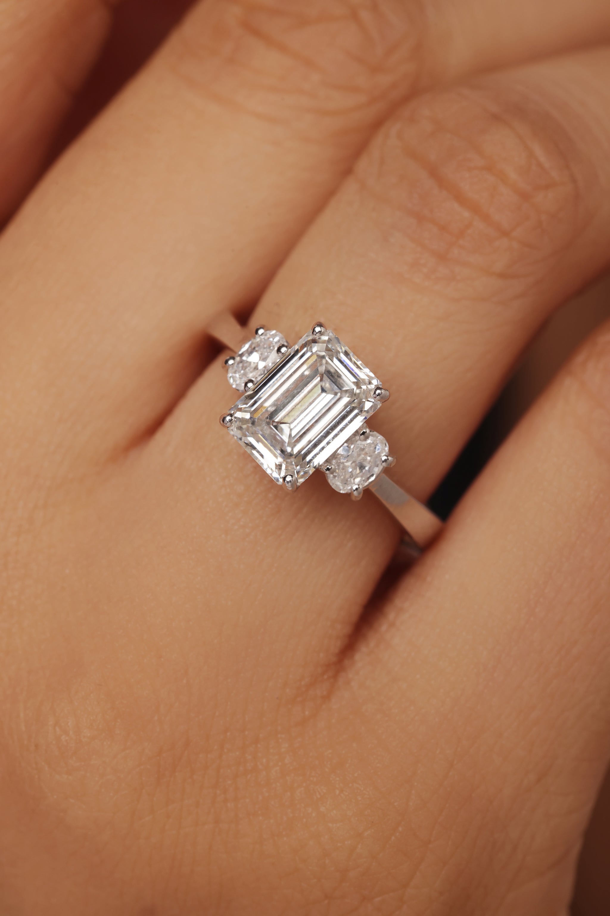 Close-up of a woman's hand showcasing a unique three-stone lab diamond engagement ring set in white gold or platinum. The ring features a rectangular emerald cut center stone flanked by two smaller oval-shaped side diamonds. This distinctive setting is ideal for promoting modern ethical fine jewellery and high-clarity diamond rings.