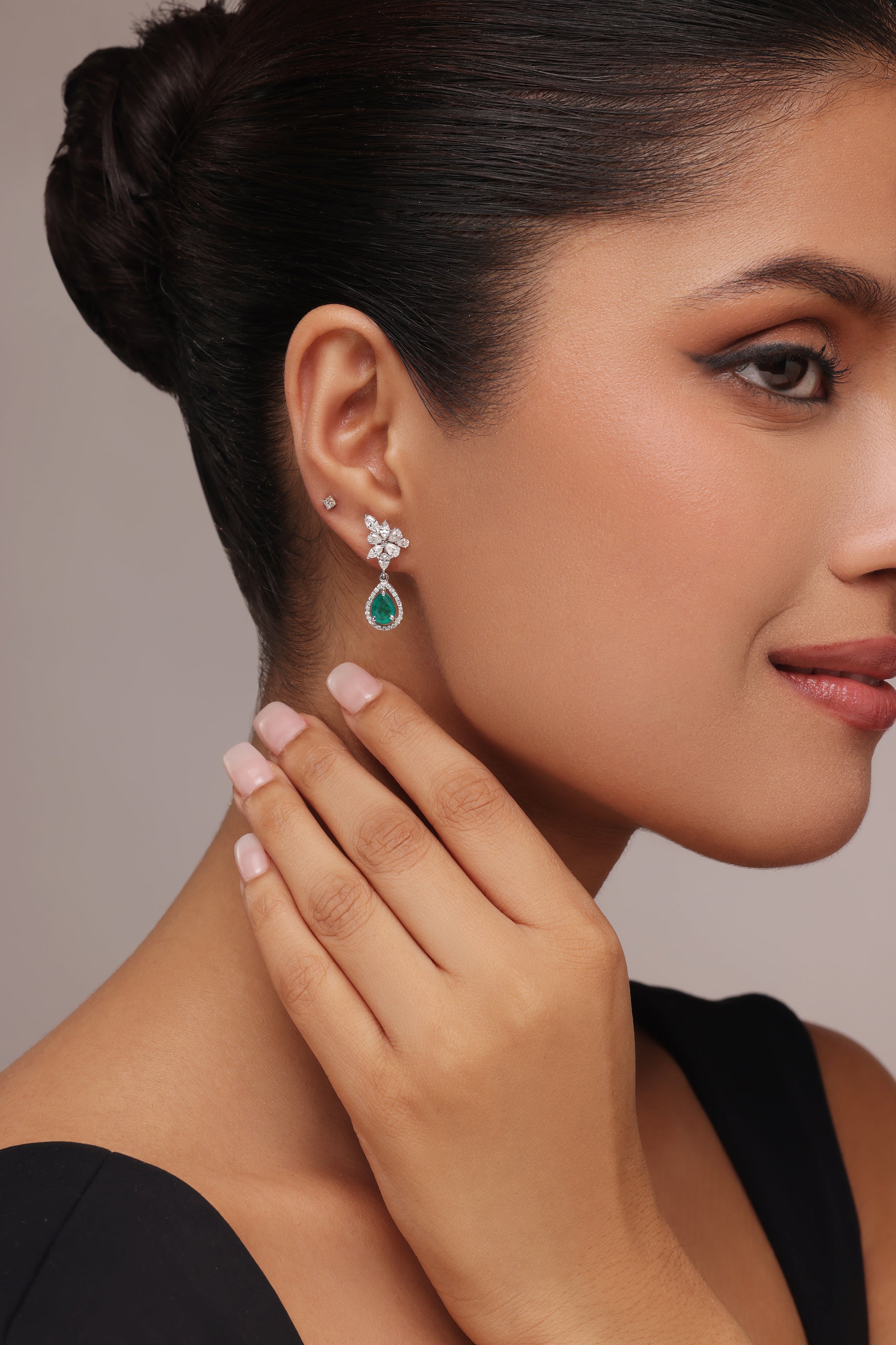 Close-up side portrait of a woman gently touching her neck, showcasing a pair of teardrop-shaped green gemstone and clear stone cluster drop earrings.