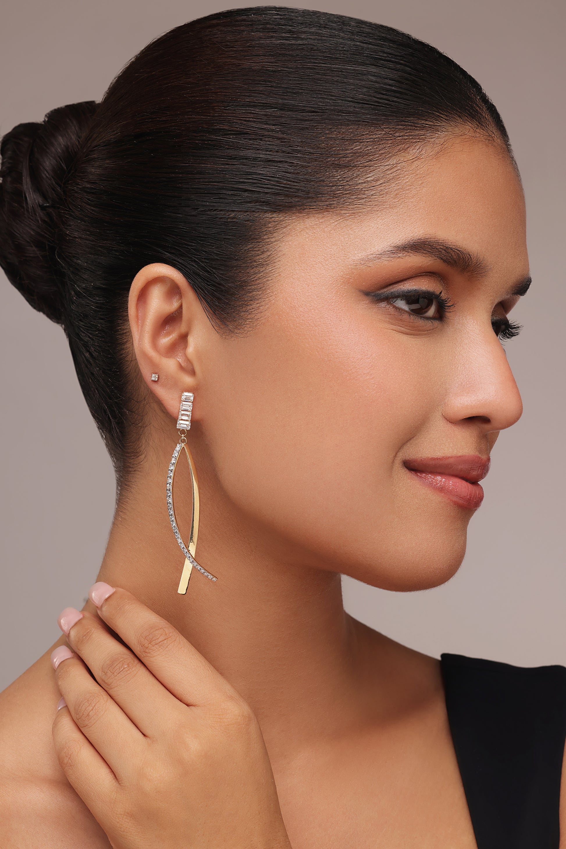 A woman in a black top showcases her ear, clearly wearing modern, asymmetrical drop earrings. The design consists of a baguette-cut stone stud attached to a long, twisting element of metal and pavé-set clear stones.