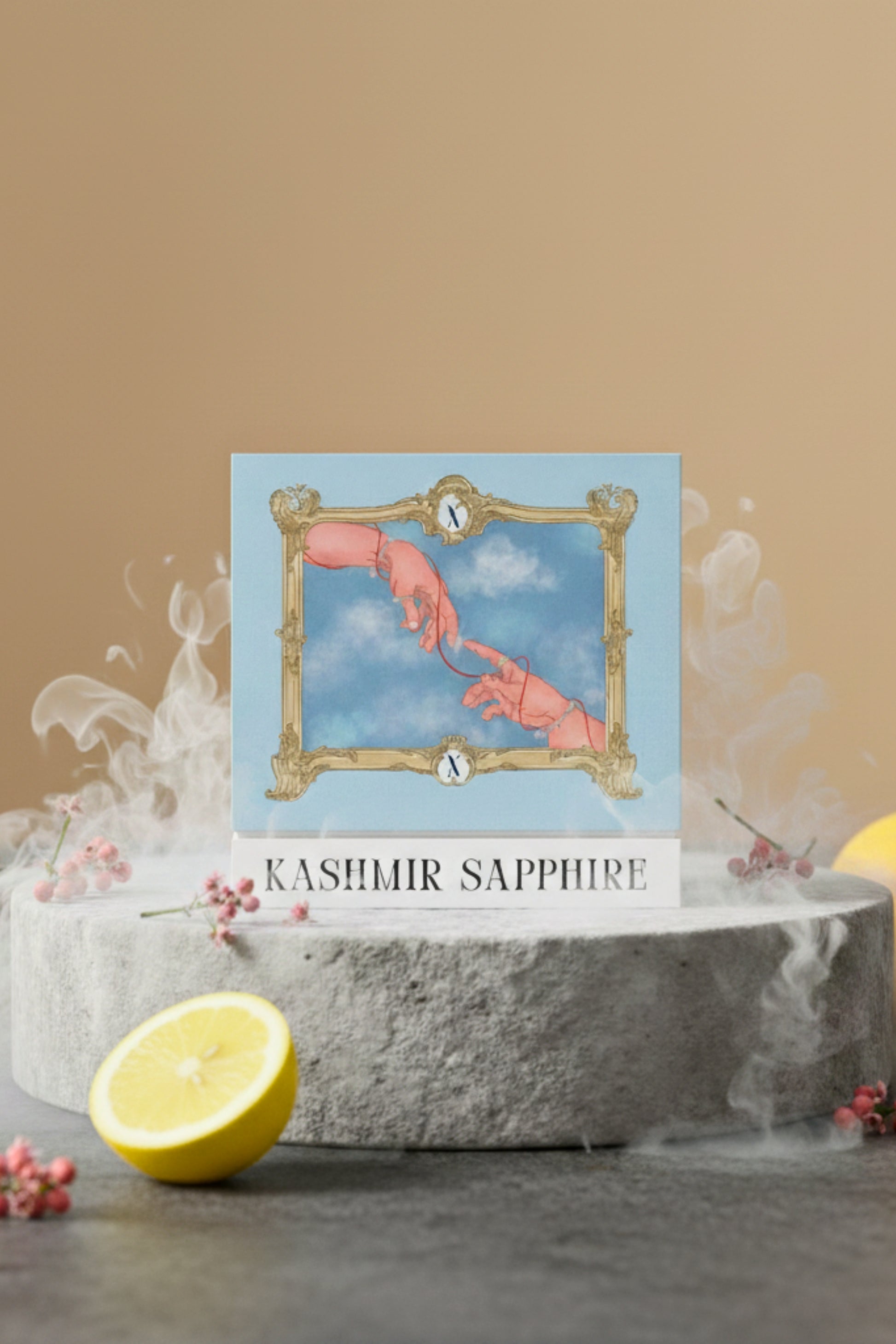 Studio product shot of the XAZINA 'Kashmir Sapphire' perfume box, featuring a blue and gold illustration of two hands reaching for each other, connected by a red string, framed by a baroque border. The box is set on a stone surrounded by a lemon slice and white mist. Represents the XAZINA brand story of connection and luxury fragrance.