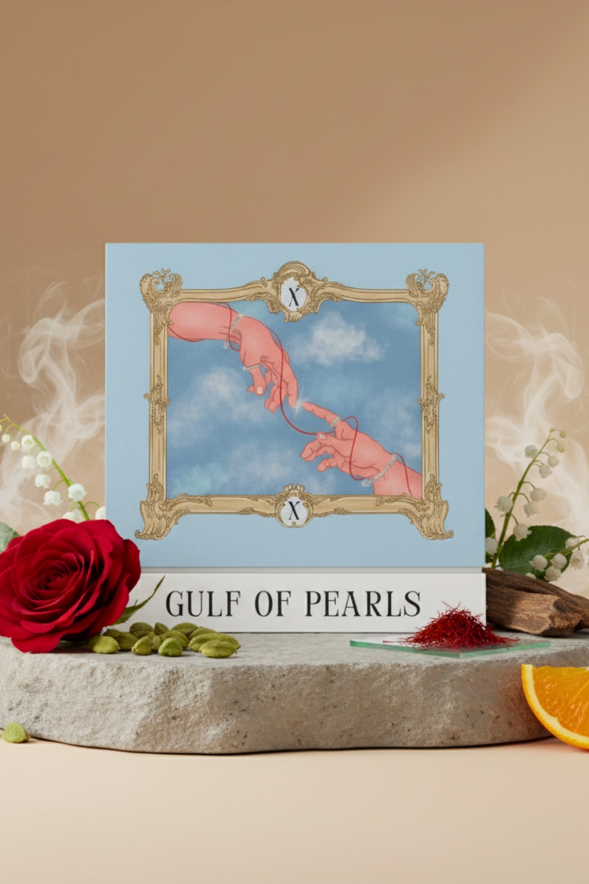 Studio product shot of the XAZINA 'Gulf of Pearls' perfume box, featuring a blue and gold illustration of two hands reaching for each other and connected by a red string, framed by a baroque border. The box is set on stone and surrounded by fragrance notes like a red rose, lily of the valley, cardamom, and saffron. Represents the XAZINA brand story of connection and luxury.