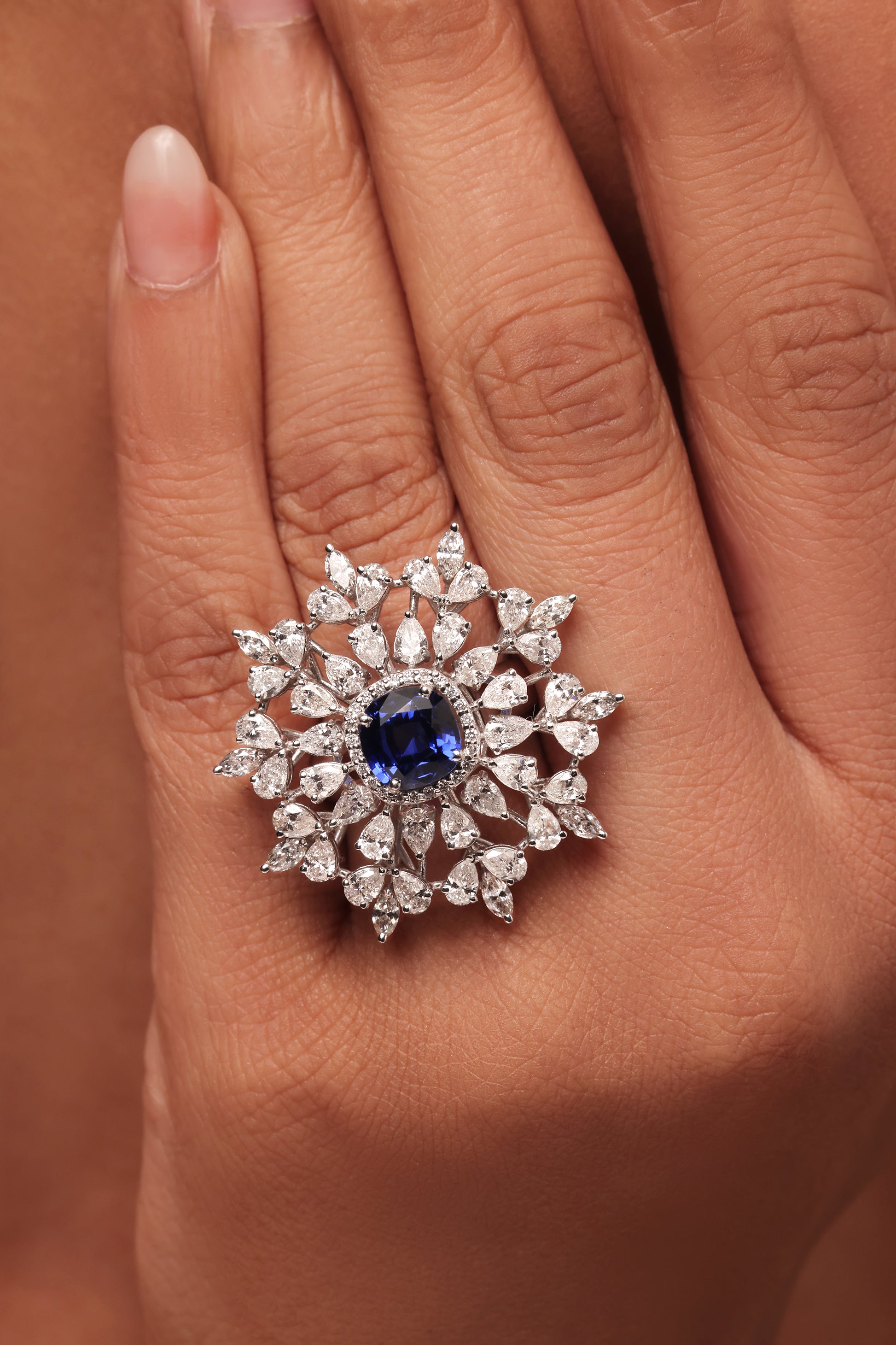 Close-up shot of a hand modeling a large cluster ring with lab grown diamond in it, featuring a central round blue stone surrounded by a dazzling burst of clear marquise and pear diamond accent stones.