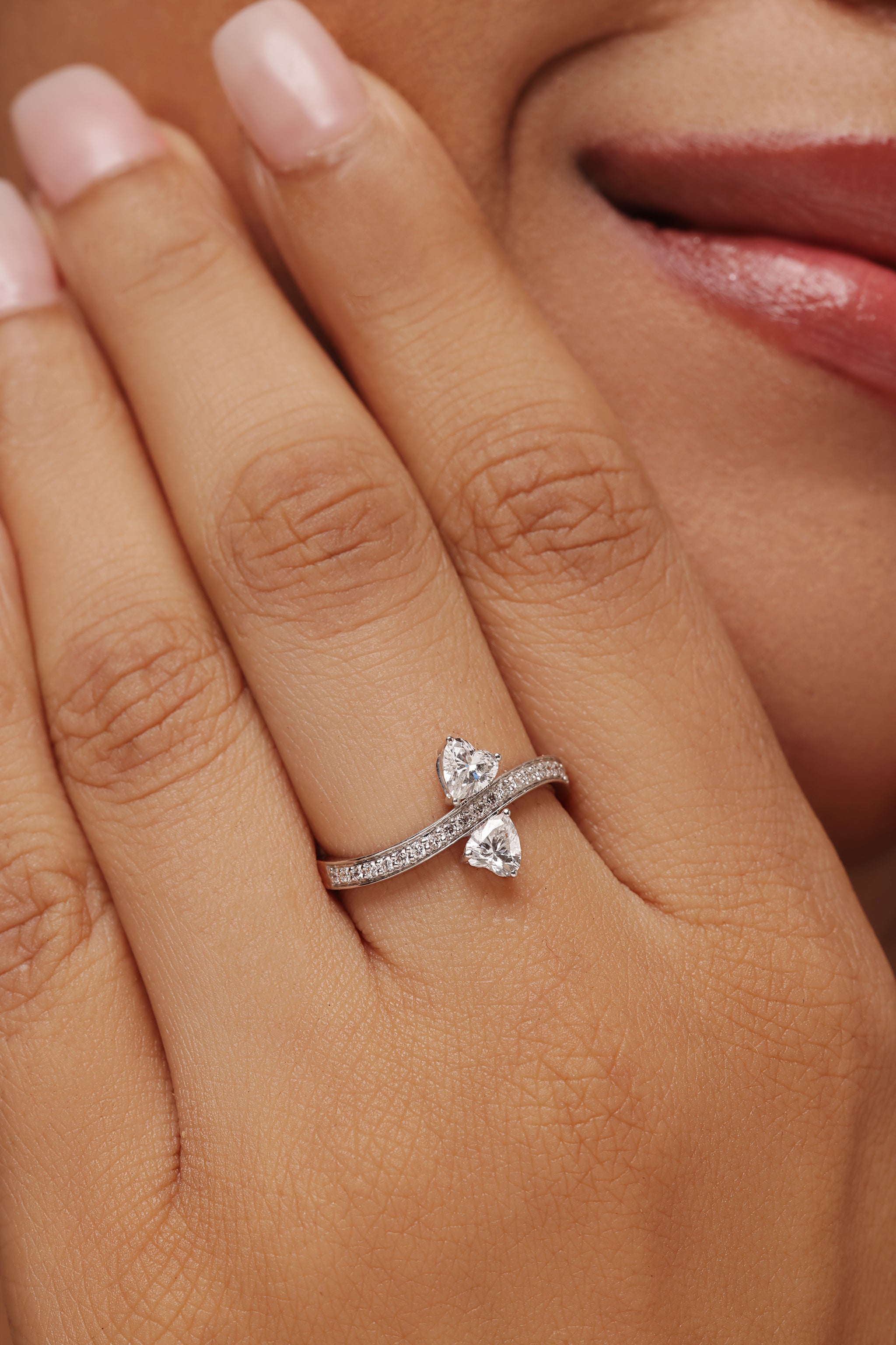 Close-up of a woman's hand showcasing a unique two-stone bypass ring set in white gold, featuring a heart-shaped and a heart lab-grown diamond in the center. The band is set with shimmering pavé diamonds. Ideal for a modern, romantic ethical engagement ring or cocktail ring.