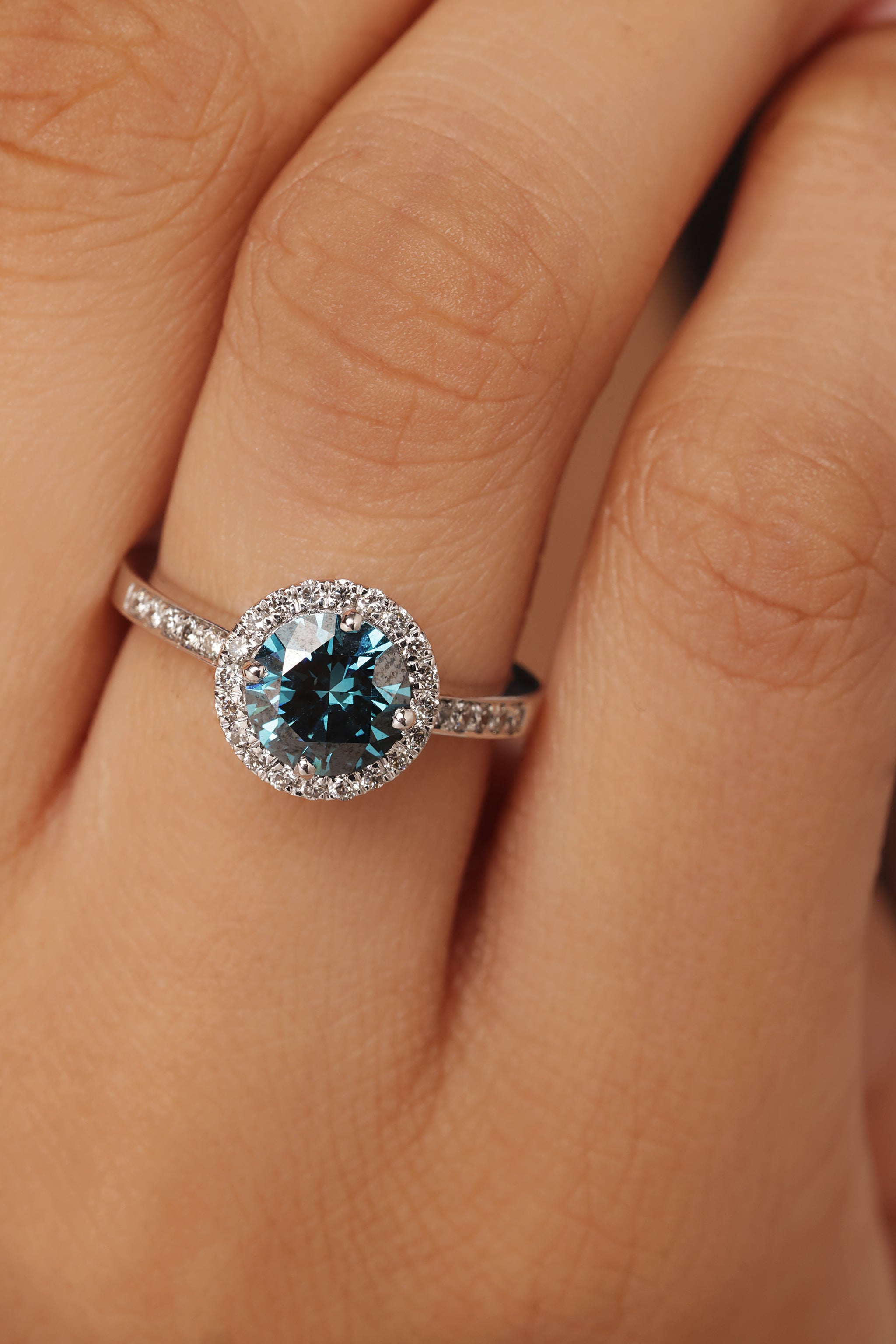 Close-up shot of a hand modeling a ring featuring a vibrant blue stone center, encircled by a round halo of lab grown diamond in it, and set on a round pave band.