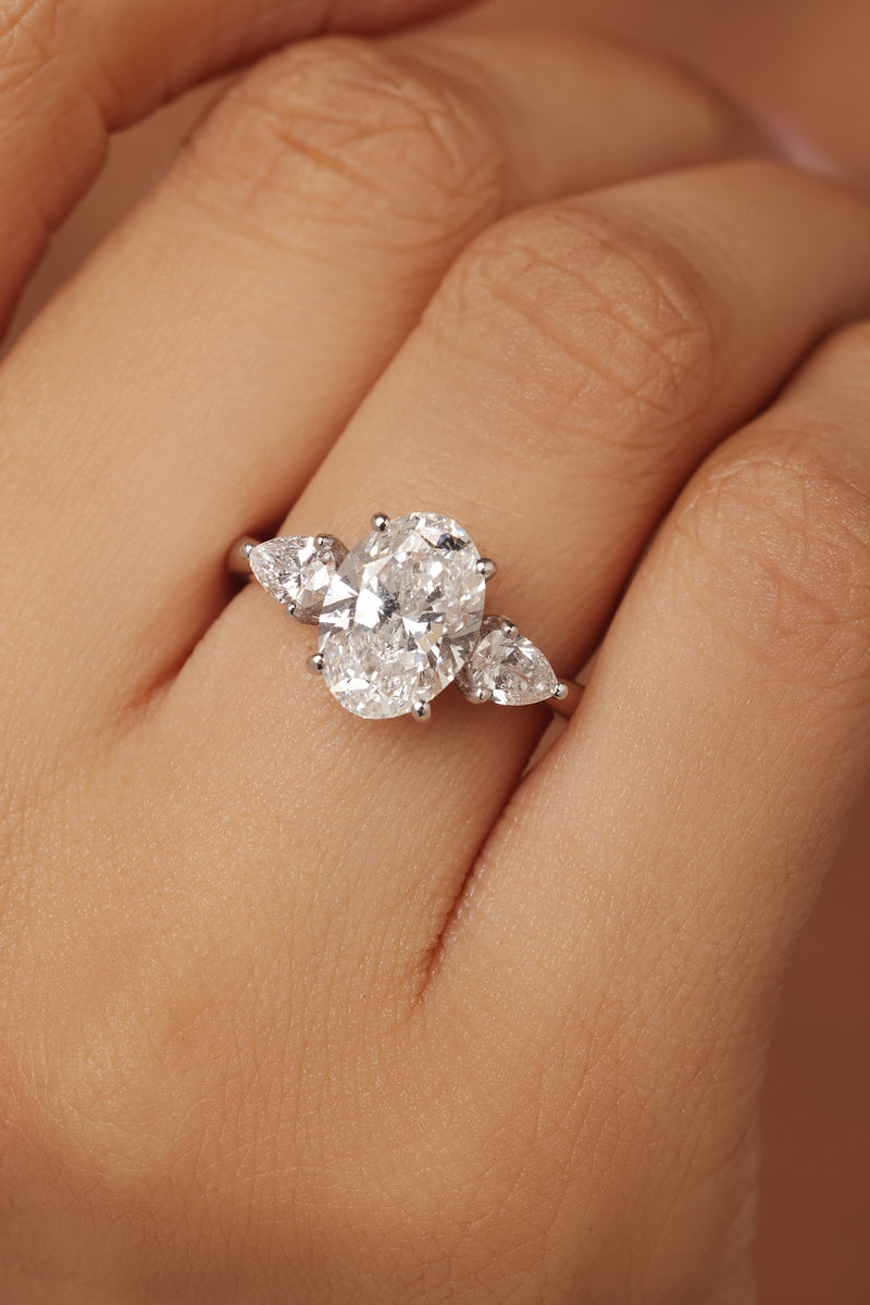 Close-up shot of a hand modeling a female diamond ring with lab grown diamond in it, featuring a prominent oval cut center stone flanked by smaller pear-shaped accent diamonds on a simple band.