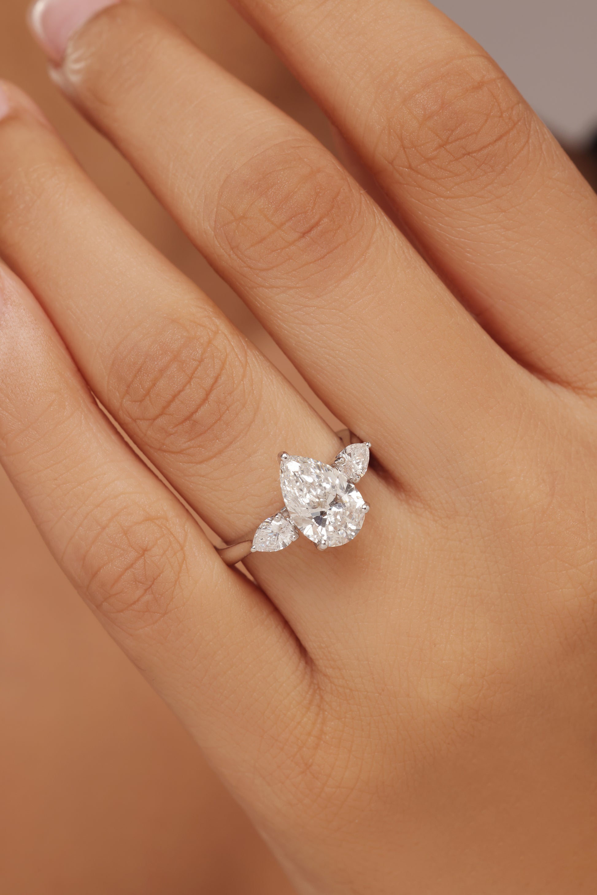 Close-up shot of a hand modeling a female diamond ring with lab grown diamond in it, featuring a prominent pear cut center stone flanked by smaller pear-shaped accent diamonds on a simple band.