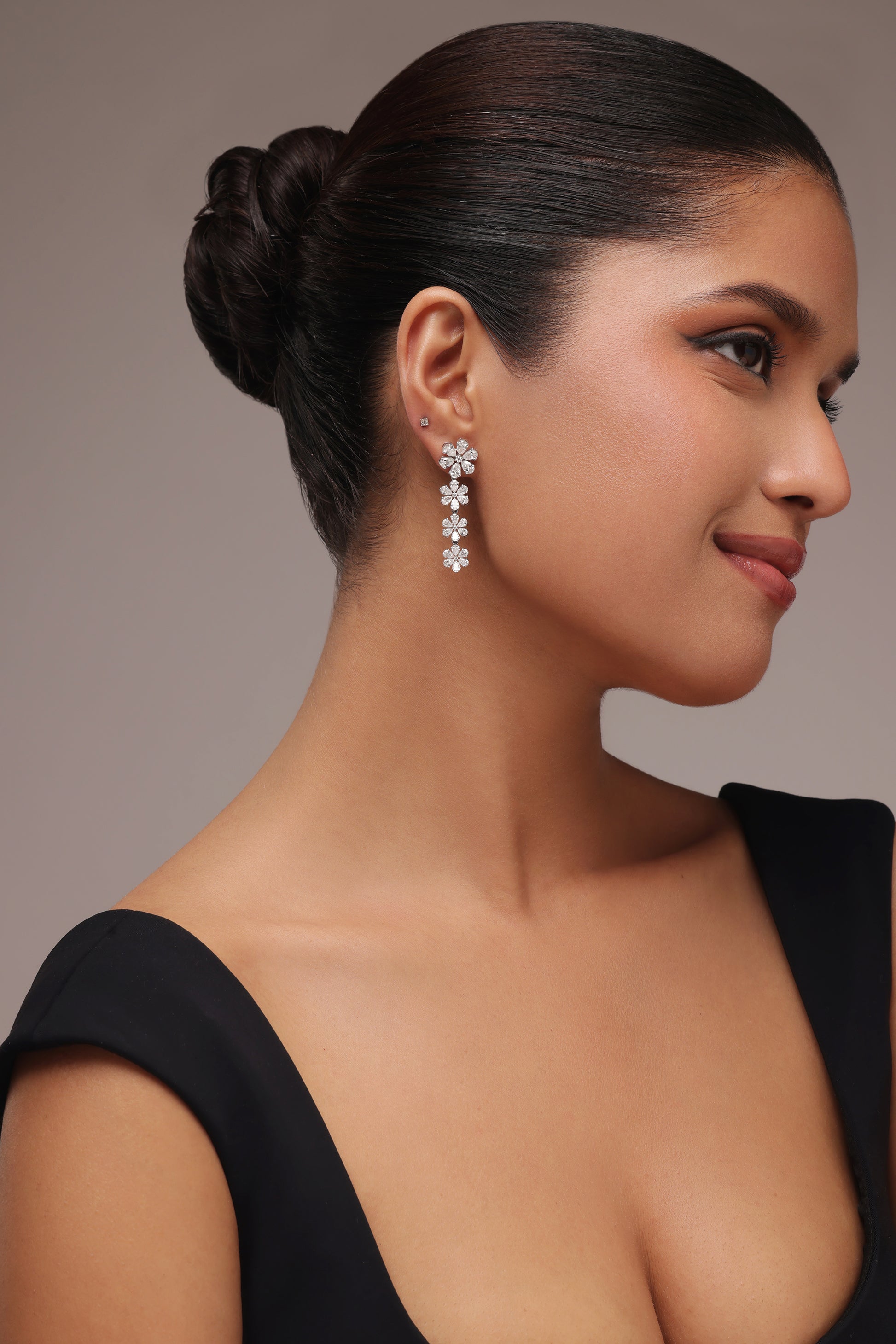 A woman in a black top with her hair pulled back showcases sparkling statement drop earrings with lab grown diamond in it, which form a delicate vertical line of four connected clear stone flower pear-cut clusters.