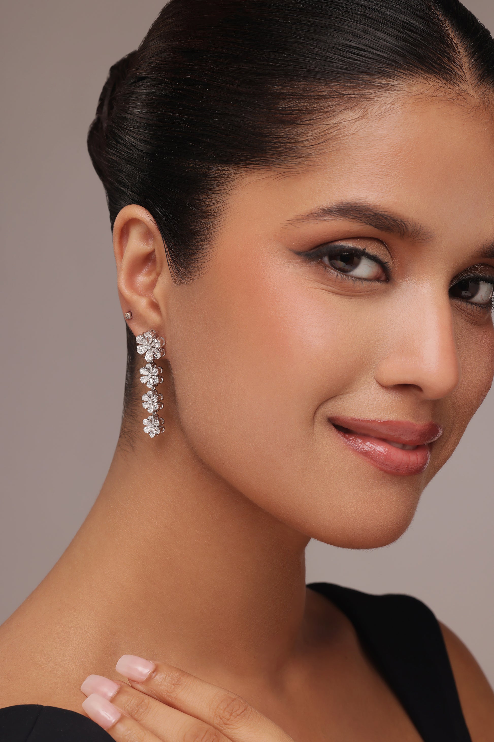 A woman in a black top with her hair pulled back models sparkling statement drop earrings with lab grown diamond in it, which form a vertical line of four clear stone floral designs.