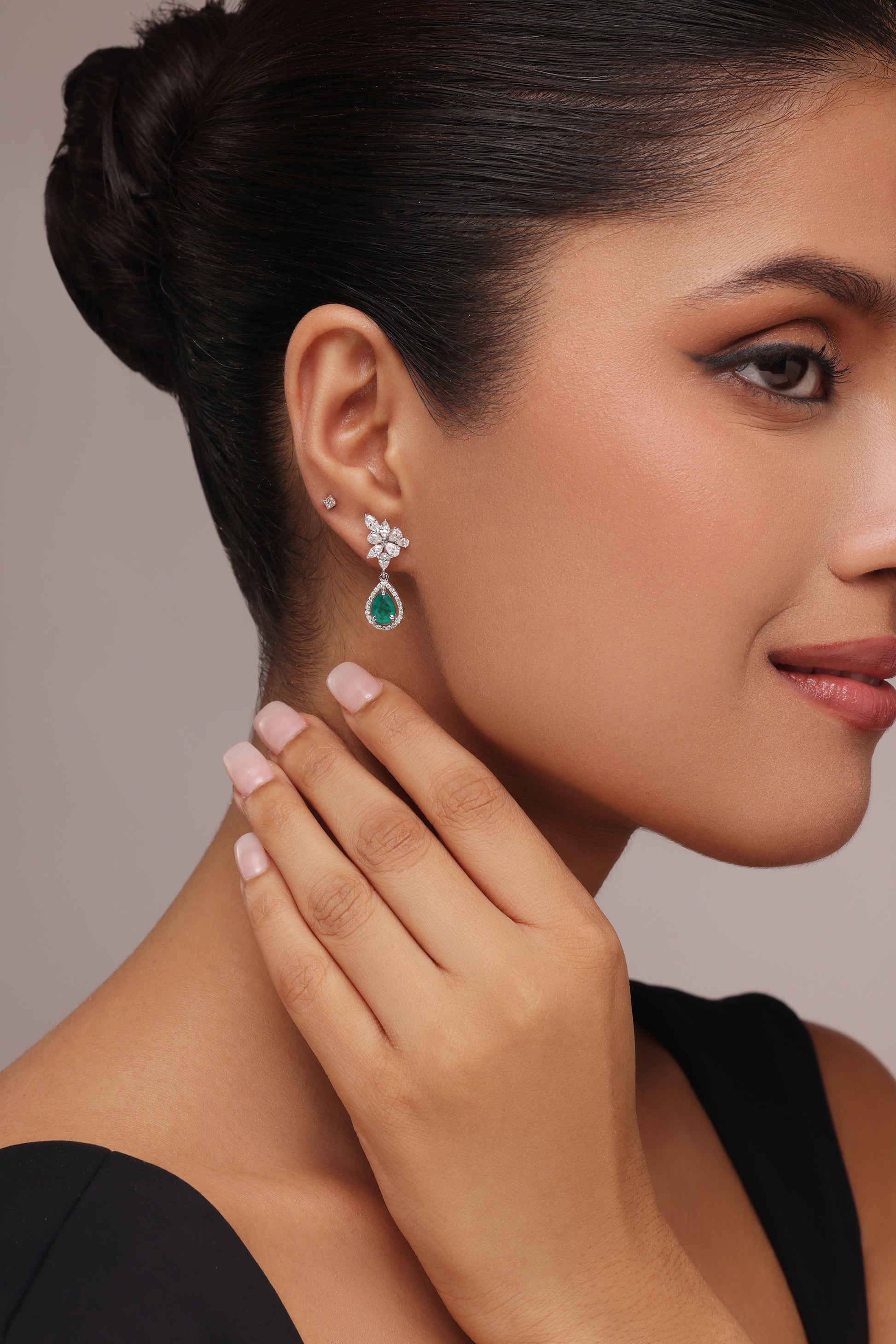 Close-up side portrait of a woman gently touching her neck, showcasing a pair of teardrop-shaped green gemstone and clear stone cluster drop earrings.