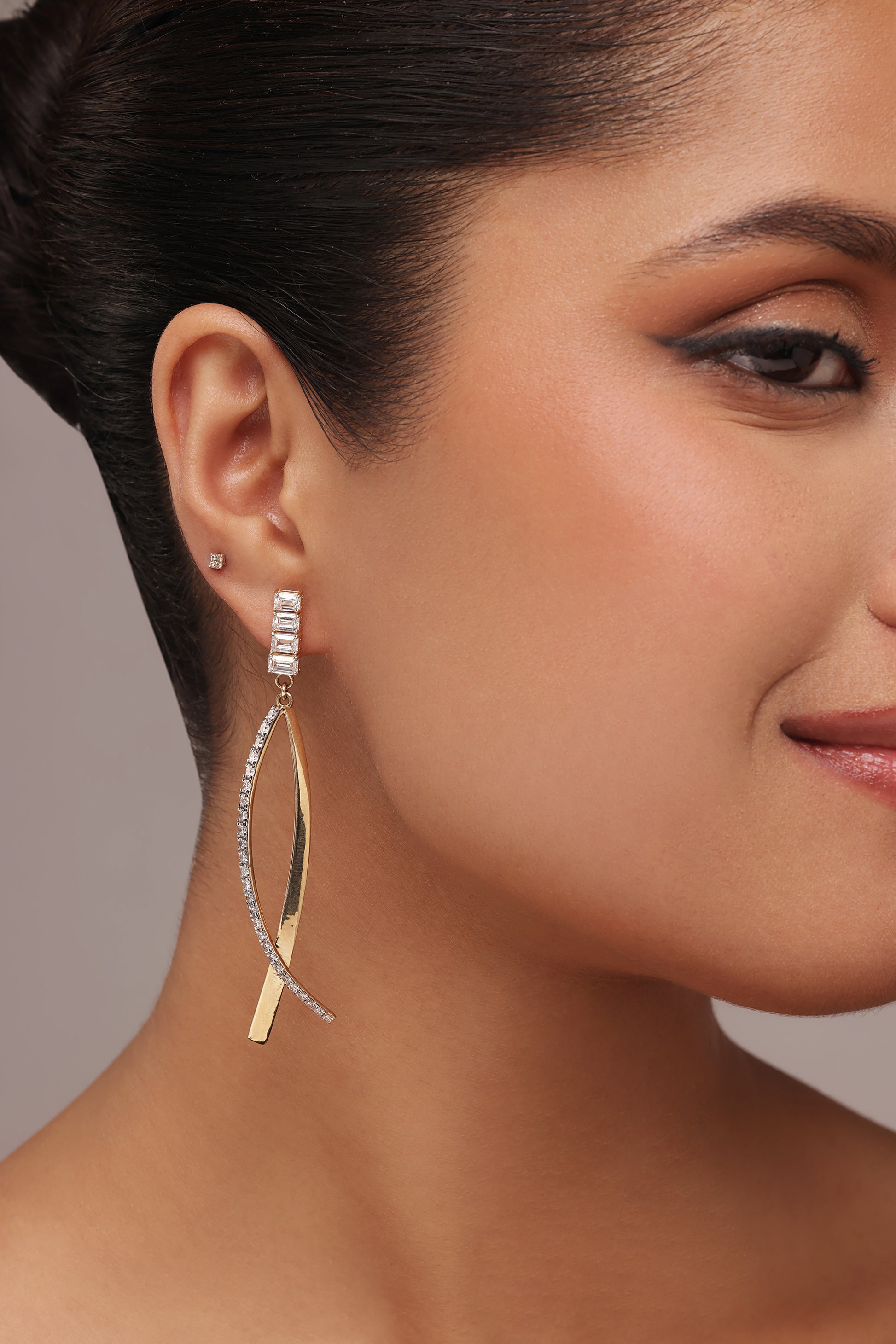 Side profile of a woman clearly displaying contemporary lab grown diamond drop earrings. The design connects a baguette-cut stone cluster stud to two long, crisscrossing elements—one plain gold-tone, the other accented with pavé-set clear stones.