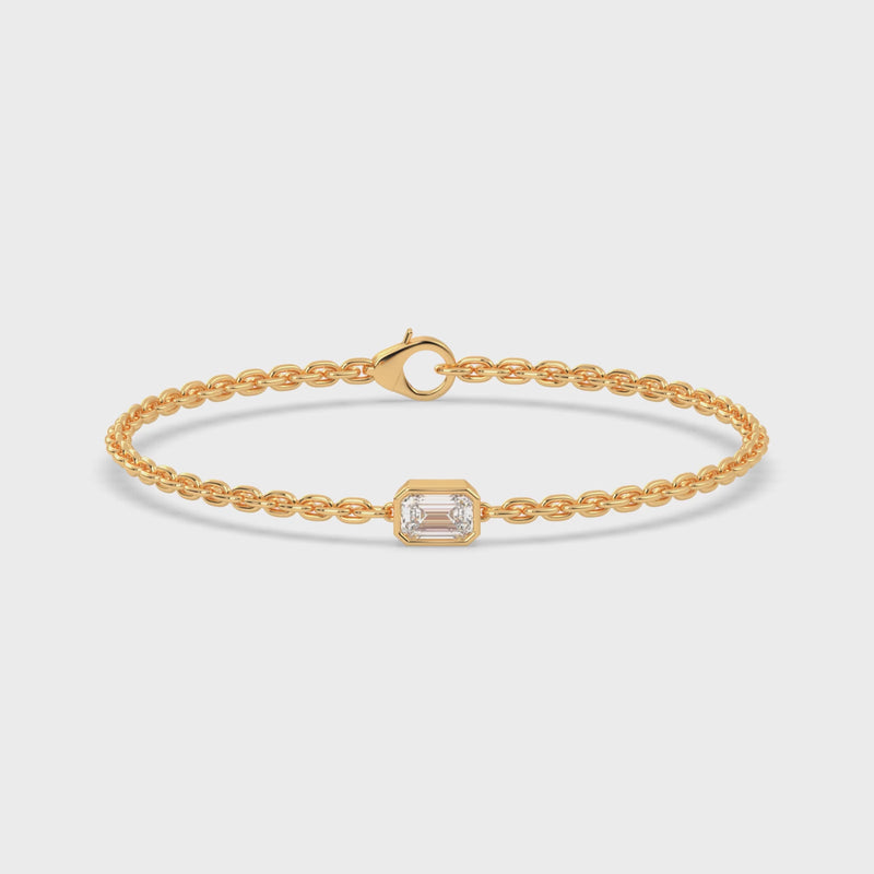 A yellow gold chain bracelet with a single, horizontally set emerald cut lab grown diamond in it at the center, secured by a clasp.
